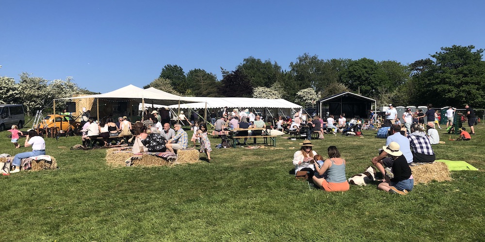 A sunny outdoor beer festival at Ditchling Camping with people sitting on hay bales and benches under large white marquees in a green field