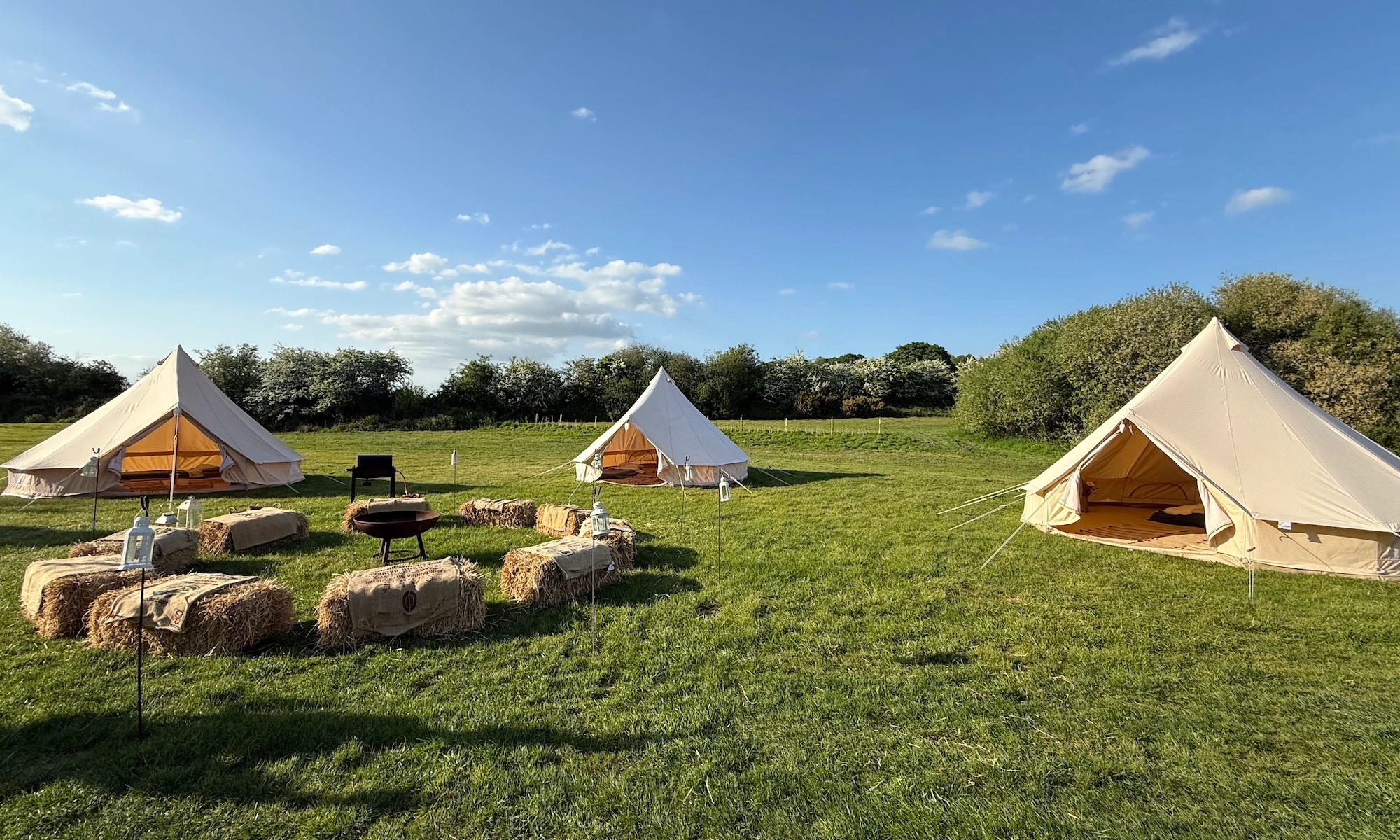 A group glamping setup at Ditchling Camping featuring three cream bell tents arranged around a central fire pit with hay bale seating under a blue sky