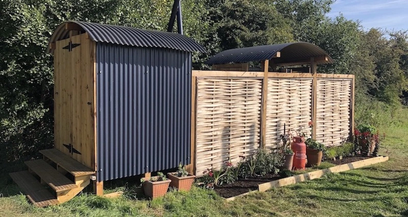 Rustic outdoor hot shower facilities with wooden cladding and flower beds at a Sussex campsite.
