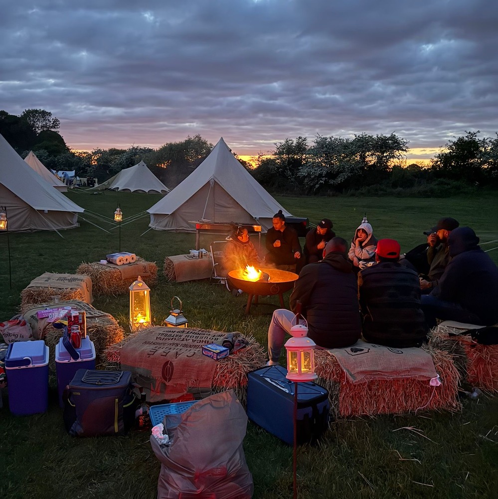 Group of friends sitting on hay bales around a campfire at Ditchling Camping glamping site with bell tents at dusk