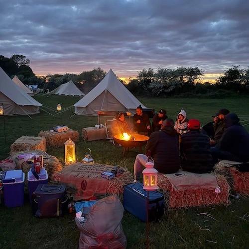 Group of friends sitting on hay bales around a campfire at Ditchling Camping glamping site with bell tents at dusk