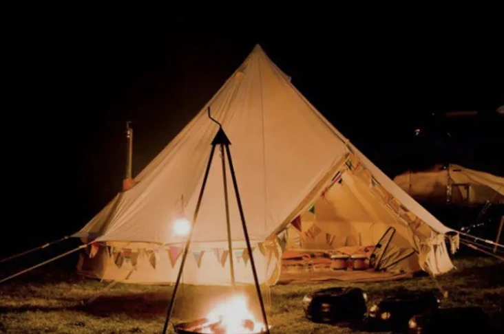 Luxury bell tent glamping at night with a glowing interior, colorful bunting, and a cozy campfire in the foreground at Ditchling Camping.