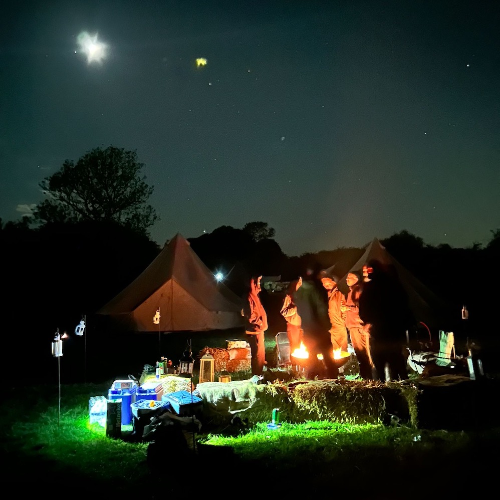 Stargazing at Ditchling Camping with people gathered around a campfire near glamping bell tents at night.